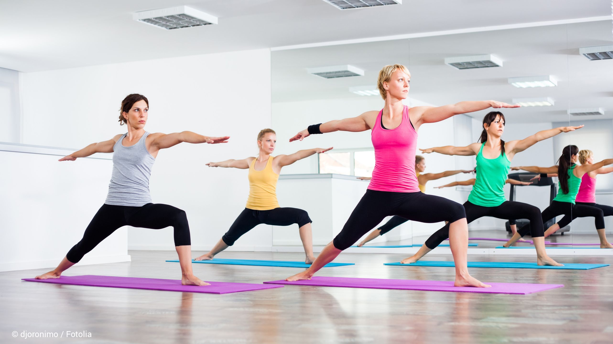 Frauen stehen in einer aufrechten Yoga-Position mit einem Ausfallschritt und ausgestreckten Armen auf Yogamatten in einem Kursraum mit einem Spiel im Hintergrund