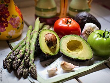 Gesunder Gemüseteller bestehend aus einer aufgeschnittenen Avocado, zwei ganze Tomaten in rot und grün, grünem Spargel und zwei Knoblauchzehen